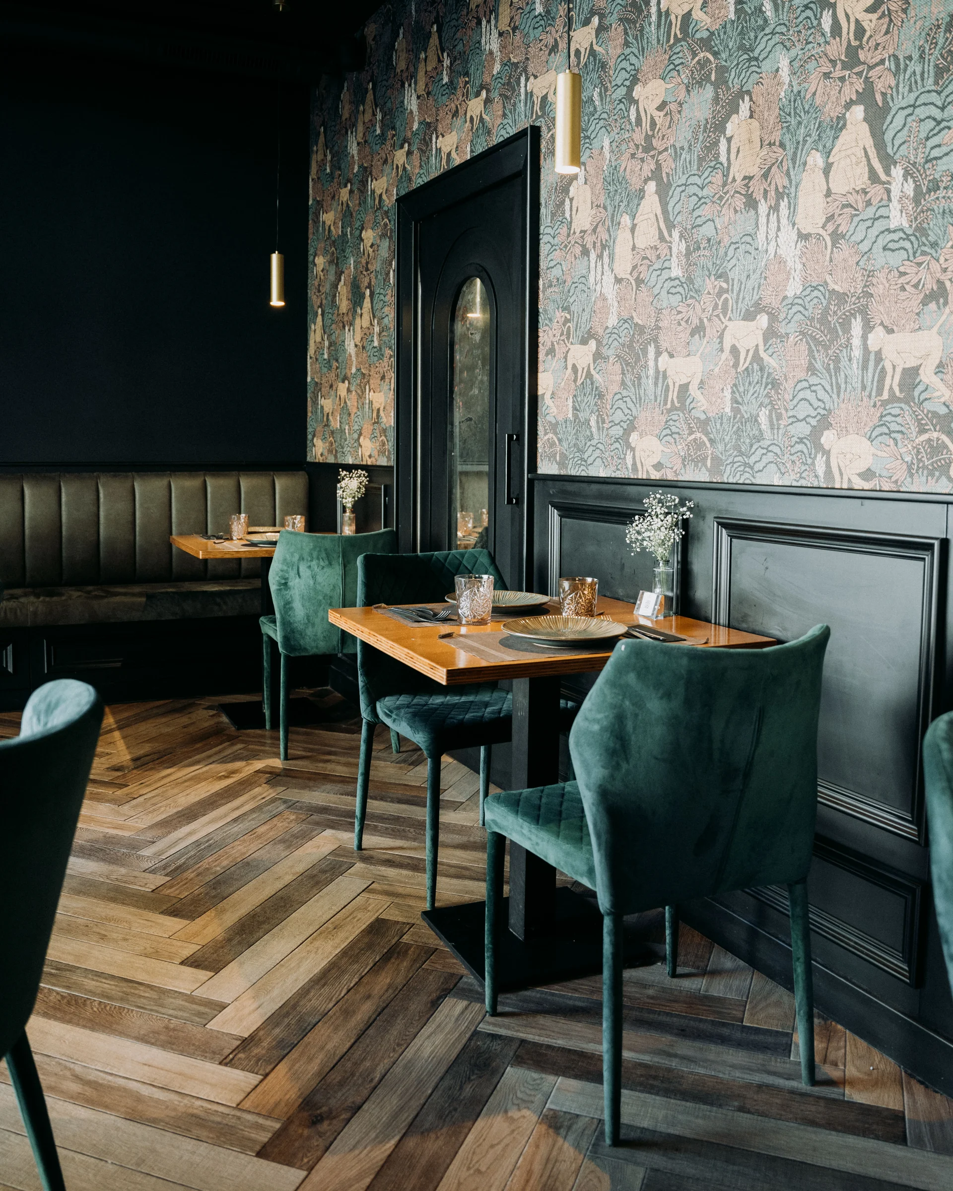 Comedor con suelo y mesas de madera, asientos de terciopelo y pared con estampado selvático