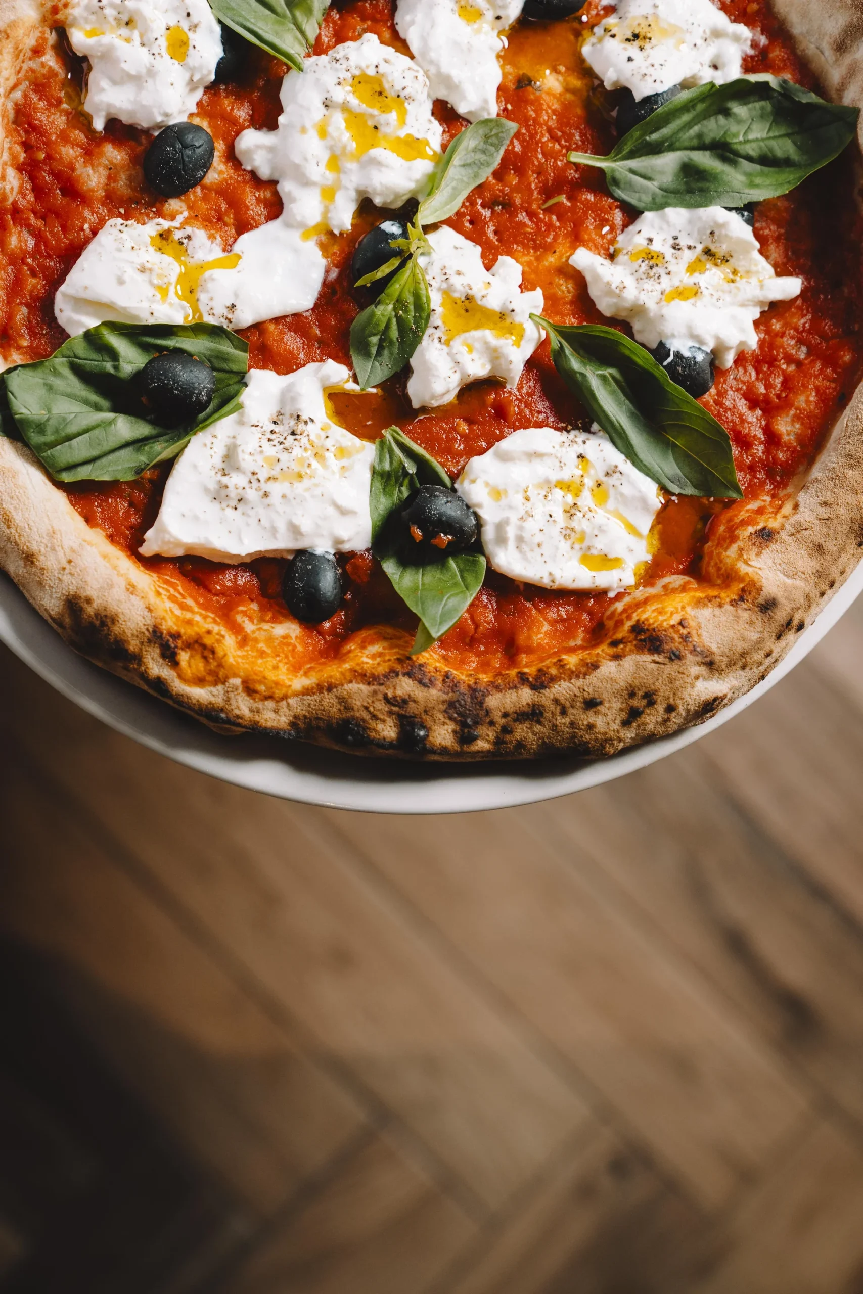 Vista superior de pizza de base roja con abundantes porciones de queso burrata fresco, aceitunas negras y hojas de albahaca, viéndose por debajo el suelo de madera rústica difuminado.